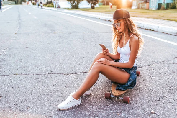 Seorang gadis dengan topi baseball duduk di atas skateboard, papan seluncur. Di musim panas di kota dengan latar belakang jalan aspal, seorang wanita muda. Rambut panjang dengan kacamata hitam. Ruang kosong untuk teks . — Stok Foto