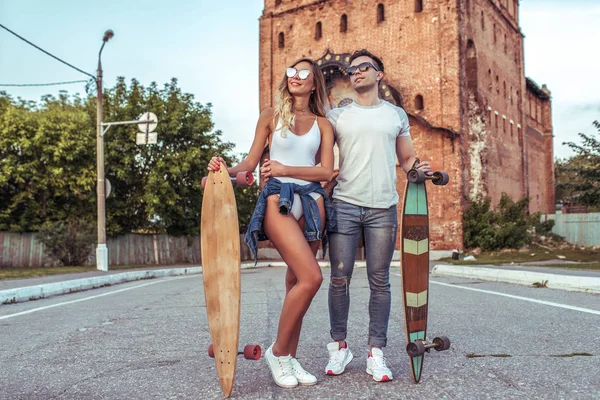 Muda dan cantik pasangan pria dan wanita, di musim panas di kota berdiri di jalan, di tangan papan seluncur dan skating longboard. Dengan pakaian biasa dan kacamata hitam. Ruang kosong untuk teks . — Stok Foto