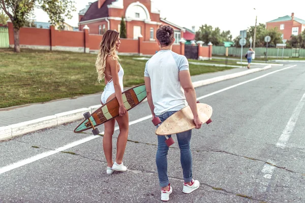 Młoda para mężczyzna i Kobieta, latem w mieście przejdź na drodze, w rękach planszy longboard i jazda na deskorolce. W ubranie i słoneczny widok od tyłu. Podróży w mieście nowe letnie. — Zdjęcie stockowe