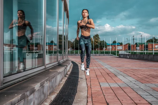 Chica atleta, ciudad de verano. Escucha música en los auriculares, salta en la carrera nocturna. En zapatillas de leggings. Mujer con tatuajes. Espacio libre para texto. En manos de un smartphone . — Foto de Stock