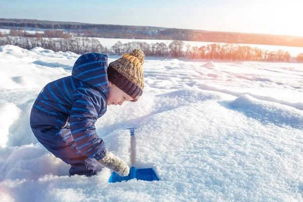 Ein kleiner Junge spielt im Winter im Schnee. Freiraum für Text. in blauen Overalls und Hut. Blick auf eine Schaufel in einer Schneewehe. — Stockfoto