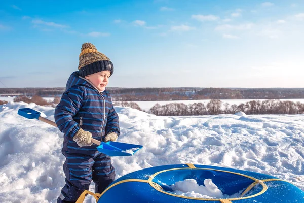 Ένα μικρό παιδί αγόρι παίζει στο χιόνι το χειμώνα. Ελεύθερος χώρος για το κείμενο. Με τις μπλε φόρμες και καπέλο. Στη λεπίδα φέρνει χιόνι στη σωλήνωση. — Φωτογραφία Αρχείου