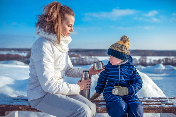 Ibu muda menuangkan secangkir teh dari termos. Anak kecil itu melihat mug. Di musim dingin, di kota di bangku di halaman bersalju . — Stok Foto