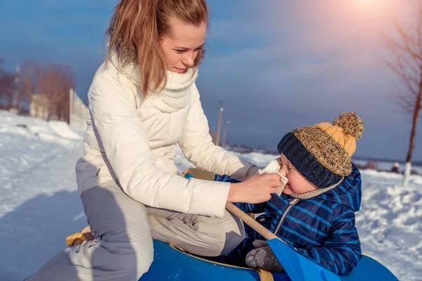 Jonge vrouw moeder veegt haar neus voor kind, een jongen, een servet. In de winter in stadspark op straat. Ouderlijke zorg voor kinderen. Het concept van de ziekte en koud en runny neus bij kinderen. — Stockfoto