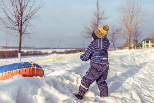 En liten pojke på vintern i staden, i en blå overall och en varm gul hatt. I händerna på att dra slang. Vila i naturen utanför staden. — Stockfoto