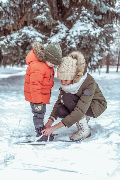 一个年轻的母亲, 一个女人为一个2-3岁的男孩系滑雪架。冬天在外面的公园里。照顾孩子, 抚养孩子。戴着温暖的帽子和夹克. — 图库照片