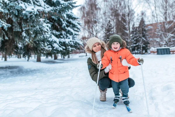 Молодая счастливая мать, женщина держит мальчика 3-5 лет сына, учится кататься на лыжах. Зимой на улице парк. Свободное место для текста. Заботиться о ребенке, растить ребенка. В теплой шляпе и куртке . — стоковое фото