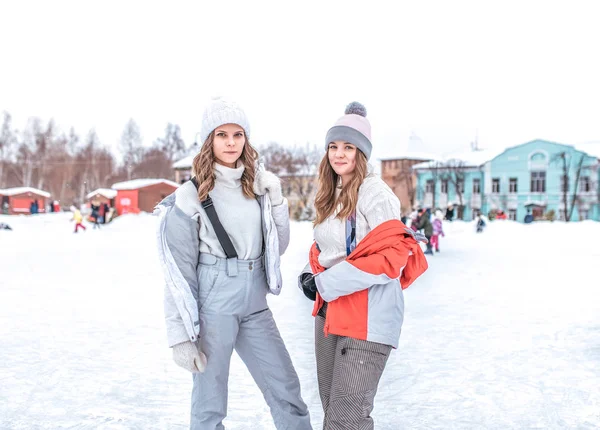 Dua gadis cantik pacar, perempuan bahagia pose tersenyum di musim dingin di kota. Dalam jaket hangat overall pakaian dan topi. Istirahat di sore musim dingin di latar belakang orang-orang skating di arena . — Stok Foto