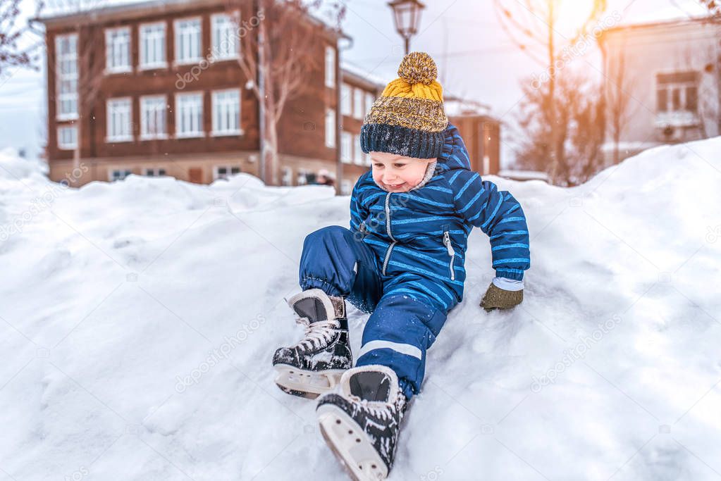 Little boy child 3-5 years old. Sits snow-covered snow drift, skates city park winter. First steps on rink. Free space for text. In blue overalls and warm hat, mittens. Happy smiling plays in snow.