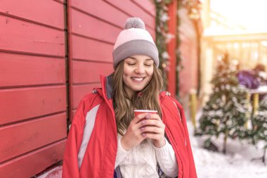 Mutlu kız Holding fincan kahve ya da çay gülümseyerek. Ahşap kepenkleri Caddesi üzerinde arka plan üzerinde kırmızı ceketli. Bir fincan sıcak bir içecek ile hakkında ısıtıyor. Bir olay olarak kışın şehirde oturan kadın.