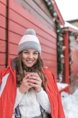 Mutlu kız duygusal holding fincan kahve gülümseyerek. Ahşap kepenkleri Caddesi üzerinde arka plan üzerinde kırmızı ceketli. Sıcak bir içecek ile fincan hakkında ısıtıyor. Bir olay olarak kışın şehirde oturan kadın.