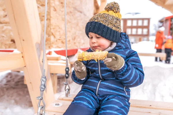 Маленький мальчик 2-6 лет, ест кукурузу в городе зимой на свежем воздухе в голубой комбинезоне шляпа и варежки. Еду на деревянных качелях. Свободное место для текста . — стоковое фото