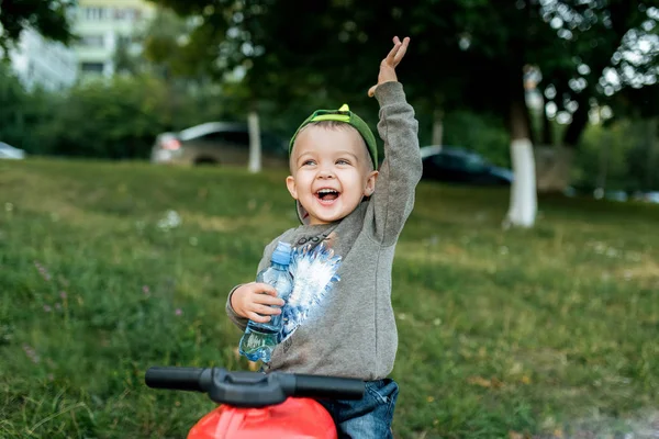 Anak laki-laki kecil berusia 3-5 tahun, duduk di atas mainan sukacita tangan ke atas, tertawa gembira di tangannya memegang botol air. Di musim panas di kota dalam udara segar . — Stok Foto