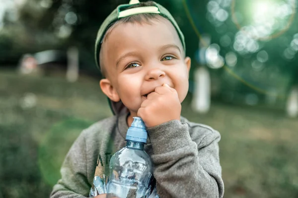 Liten pojke barn 3-5 år gammal, närbild porträtt, glad skrattande i hans händer som håller en flaska vatten. Sommaren i staden i den friska luften. En uppriktig look med ögon. — Stockfoto