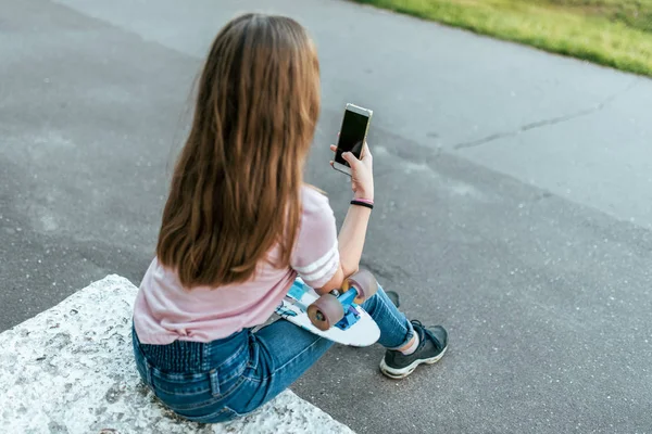 Teenie-Mädchen 9-10 Jahre alt, in den Händen eines Skates sitzend. im Sommer in der Stadt in lässigen Jeans. Kommunikation im Internet, in den Händen des Smartphones hält eine Nachricht in der Anwendung schreibt. zurück. — Stockfoto