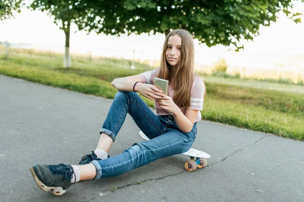 Gadis remaja 9-11 tahun, duduk di atas kapal di tangan telepon. Di musim panas di kota dengan celana jeans dan kaos merah muda. Komunikasi di Internet, di tangan memegang smartphone. Senyum dan pose . — Stok Foto