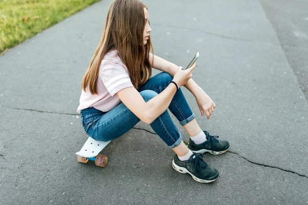 Gadis remaja 9-11 tahun, duduk di atas kapal di tangan telepon. Di musim panas di kota dengan celana jeans biasa. Komunikasi di Internet, di tangan smartphone memegang, korespondensi dalam jejaring sosial . — Stok Foto