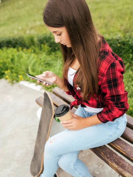 Seorang gadis remaja berusia 10-13 tahun duduk di bangku di tangannya memegang smartphone dan secangkir teh. Komunikasi di Internet melalui aplikasi. Di musim panas di kota. Di dekatnya ada skateboard . — Stok Foto