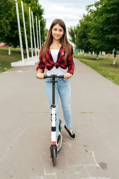 Adolescenta de 11-12 ani stă în parc pe un skateboard în timpul verii pe fundalul copacilor verzi. Fericit zâmbind călărind un scuter într-un parc de oraș . — Fotografie, imagine de stoc