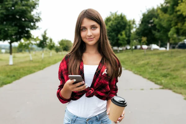 Gadis cantik dengan rambut panjang remaja 11-12 tahun, berdiri di musim panas di taman kota. Di tangan smartphone dan secangkir kopi. Komunikasi remaja dalam jejaring sosial. Selamat tersenyum . — Stok Foto
