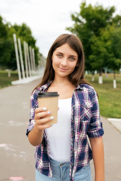 Девочка-подросток 11-12 лет, стоящая в летнем городском парке. Счастливая улыбается, протягивает руку с чашкой кофе или чая. Концепция предложения выпить. Угощения и призывы к избавлению от жажды . — стоковое фото