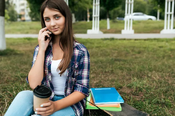 Adolescente intelligente de 11-12 ans, assise en été dans le parc de la ville. Sourires heureux, appels sur Internet. A proximité se trouvent des carnets de notes avec documents, planche à roulettes et une tasse de café ou de thé. Espace libre pour le texte . — Photo