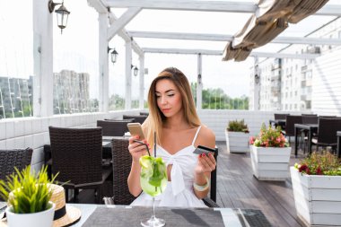 Güzel kız uzun saçlı elbiseli yaz aylarında verandada bir kafede oturuyor. Elini içinde bir plastik kart ve telefon. Bir kadın öğle veya akşam yemeği online banka ile kredi kartıyla ödeme yapar.