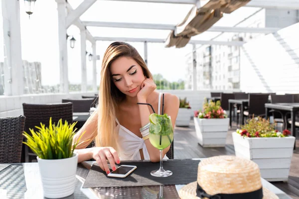 Joven y hermosa chica en un café. Sobre la mesa una copa con un cóctel de lima verde. Una mujer lee un mensaje por teléfono, en Internet para almorzar en un restaurante. En verano en el porche del sol . —  Fotos de Stock