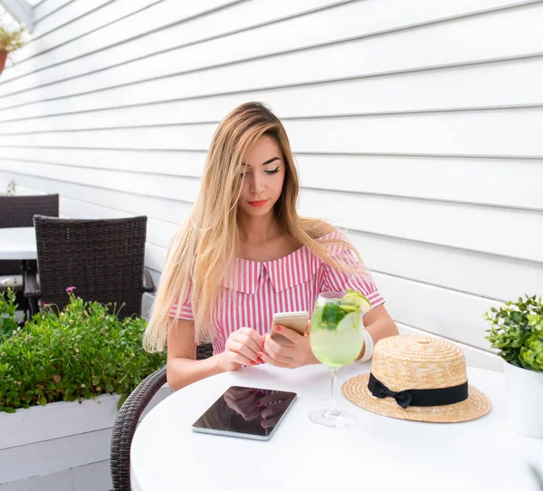 Young and beautiful girl in cafe with long hair. Reads a message on phone, online on Internet for lunch in restaurant. In summer on sun porch. On the table is a glass of green lime and a tablet.