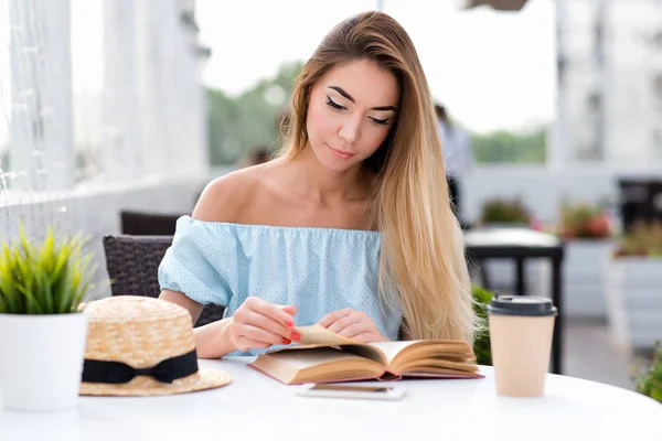 Молодая и красивая девушка с длинными волосами. На столе чашка кофе и чай. Женщина, читающая бумажную книгу на столе. Интересный роман переворачивает страницу. Летом на солнечной террасе . — стоковое фото