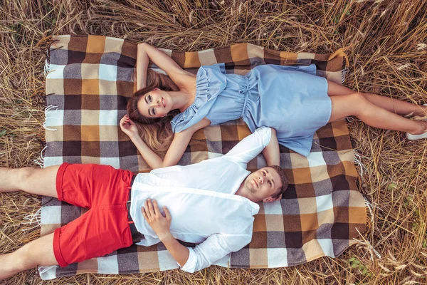 Een jong koppel, een man en een vrouw in de zomer in een tarweveld, liggen op een tapijt en ontspannen in de frisse lucht. Het concept van de liefde is vreugde en zorg in een relatie. Romantisch voldoen aan de zonsopgang en zonsondergang. — Stockfoto