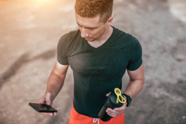 Şişe su ve telefon tutan genç bir adam. Spor zemin şehirde yaz aylarında. Uygulama Internet üzerinde eğitim denetler. Bir adam bir t-shirt ve şort sokakta yemiş kalori denetler.