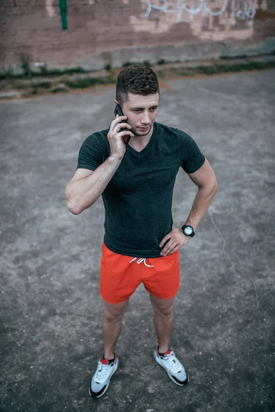 若い男は、スマート フォン上で鳴っている電話を保持しています。スポーツ グランドの都市の夏。T シャツを着た赤いショート パンツとスニーカー。新鮮な空気で運動後の休息します。. — ストック写真