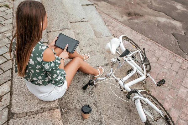 La chica se sienta en las escaleras en el verano en la ciudad. En las manos de la aplicación de tableta está abierto en Internet. Cerca de la bicicleta y una taza de café o té . —  Fotos de Stock
