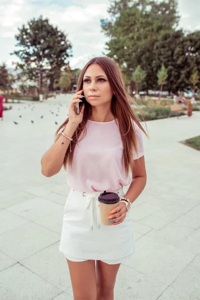 Une fille se tient en été en ville. Dans sa main, le smartphone appelle au téléphone avec une tasse de thé ou de café. Femme en vêtements décontractés . — Photo