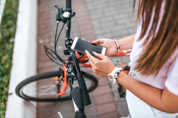Una chica está de pie con una bicicleta en el verano en la ciudad. En manos de un smartphone, escribe un mensaje a través de la aplicación en las redes sociales. Concepto de ruta de búsqueda o mapa en la ciudad . —  Fotos de Stock