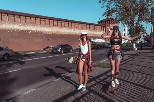 Mooie meisjes vriendinnen in zomer stad. Lopen rit longboard skateboard. Met lange haren en baseball caps zijn plezier spelen gelukkig lachend. Vrije ruimte voor tekst. Op de achtergrond van de weg met auto 's. — Stockfoto