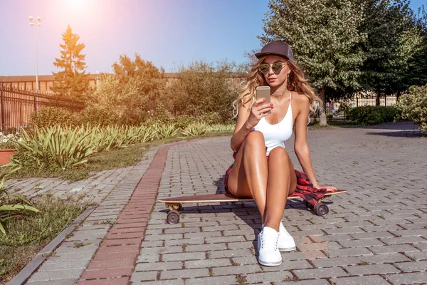 Schöne Mädchen im Sommer Stadt. sitzt auf einem Longboard-Skateboard. Mit langen Haaren und Baseballmütze schreibt er eine Nachricht ins Internet, online in sozialen Netzwerken. Freiraum für Text. — Stockfoto