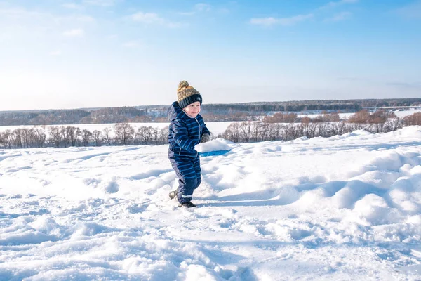 Anak kecil dengan sekop salju di musim dingin di taman. Happy membawa salju di atas sekop dalam pakaian kasual . — Stok Foto