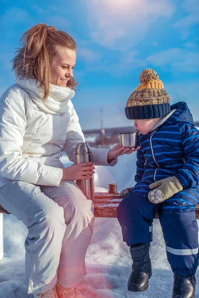 A young mother is holding a thermos with tea, a little son is blowing hot tea nearby. A woman gives a mug to child. In winter in the park on the bench.
