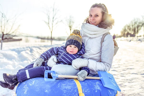 Счастливые мама и малыш сидят на трубчатой игрушке. Зимой они отдыхают на свежем воздухе, счастливо улыбаясь в руках снежной лопаты . — стоковое фото