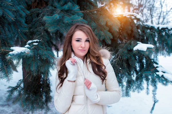 Uma menina bonita fecha seu casaco de inverno quente branco. No fundo de uma árvore coberta de neve. Uma mulher morena de luvas brancas está sorrindo . — Fotografia de Stock