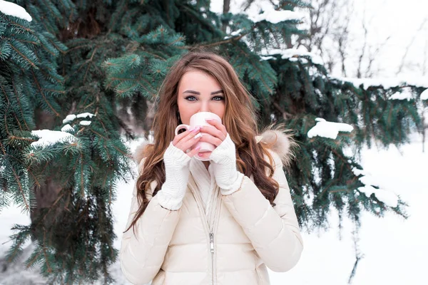 Chica morena en el bosque de invierno, en el fondo de los abetos. En sus manos sosteniendo una taza, bebiendo café caliente o té. Con una chaqueta blanca y mangas calientes. Descanse fuera de la ciudad en el bosque . —  Fotos de Stock