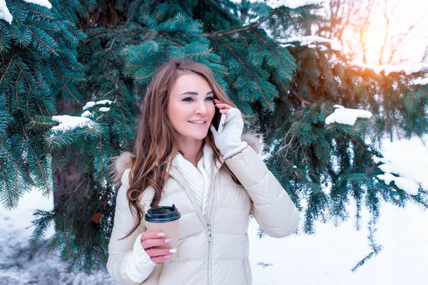 Ragazza bruna nella foresta invernale, sullo sfondo di abeti. Chiamate al telefono online da uno smartphone che parla. Nelle mani di tenere tazza. In giacca bianca e maniche calde. Riposo fuori città nella foresta . — Foto Stock