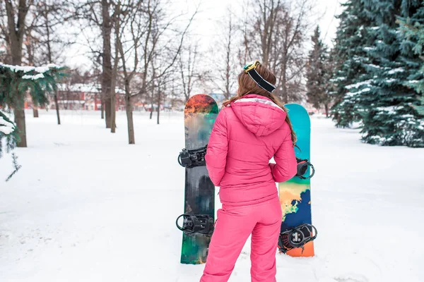 A girl in a pink jumpsuit in the winter on a forest in the forest rests. View from back on the background of snowboards in the snow. Free space for text. — Stock Photo, Image