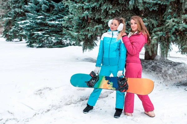 Two girls girlfriends beautiful women happy in hand of a snowboard. Resting resort, nature winter against backdrop Christmas tree. Free space for text. In sportswear pink and blue fitness meeting. — Stock Photo, Image