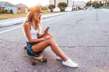 Sarışın kız güzel mayo gövdeli longboard kurulu. Yaz gün şehir. El smartphone, yazar mesaj sosyal ağ, online app Internet. Tabaklanmış deri kapağı, uzun saçlı. Boş alan.