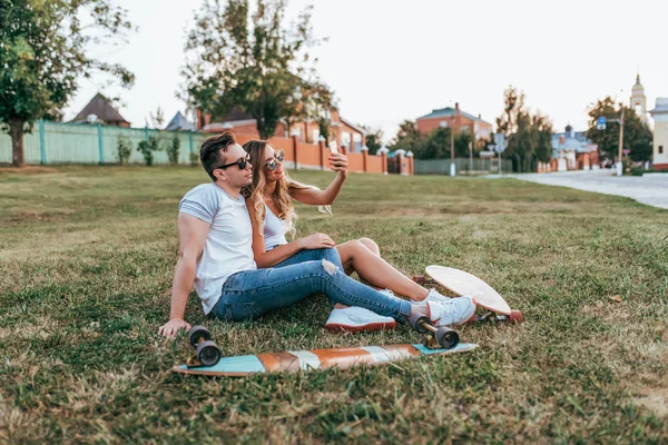 Beautiful young couple man woman, sit on green lawn summer. In hands of phone, picture selfie, board of longboard. Happy relax active lifestyle. Concept happy love and relationship, young family. — Stock Photo, Image