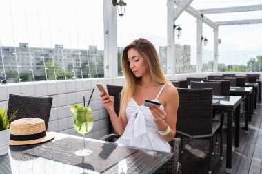 Yaz kafede gülümseyen güzel kız. Plastik kredi kartı elinde, Kahvaltı ve öğle yemeği app internet için ödeme yaparsınız. Temassız ödeme smartphone. Yazar PIN kodu açık havada kartından.