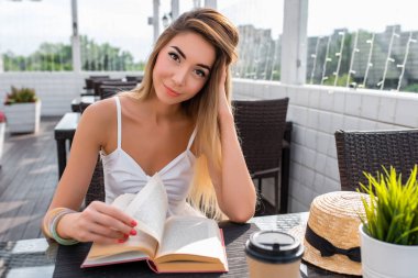 Güzel kız yaz kafede. Kağıt kitap, sanat Dedektif okur. Kahve veya çay ve bir hasır şapka, Kahvaltı ve öğle yemeği beyaz elbisenin içinde geçerli. Verandada kafede zor bir günün ardından dinlenmek.